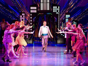 An actor in a white tuxedo struts down center stage surrounded by other dancers in formal attire on stage of broadway musical The Prom