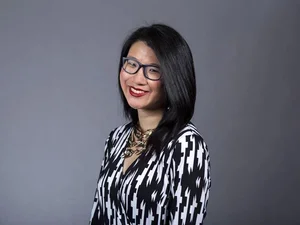 Dark haired woman wearing glasses and black and white blouse against grey background