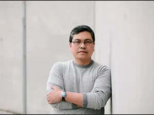 man in a striped shirt leans against a white exterior wall