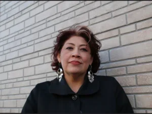 headshot of a woman in a black coat with dangling silver earrings against a white brick wall 