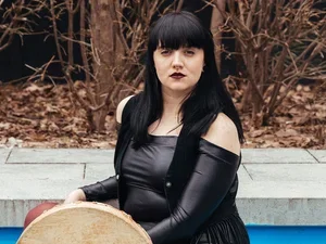 woman with black hair and a shiny dress holds a skin drum in what looks like a swimming pool