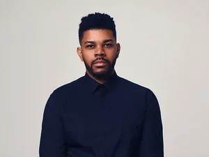 A serious-looking man with brown skin, dark hair, who is wearing a dark blue button up shirt.