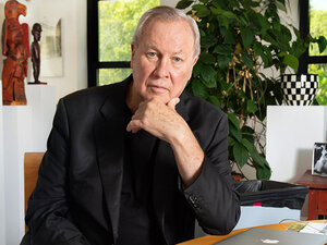 Robert Wilson, director and artist, seated at a desk with sculptures and plants in a well-lit room.