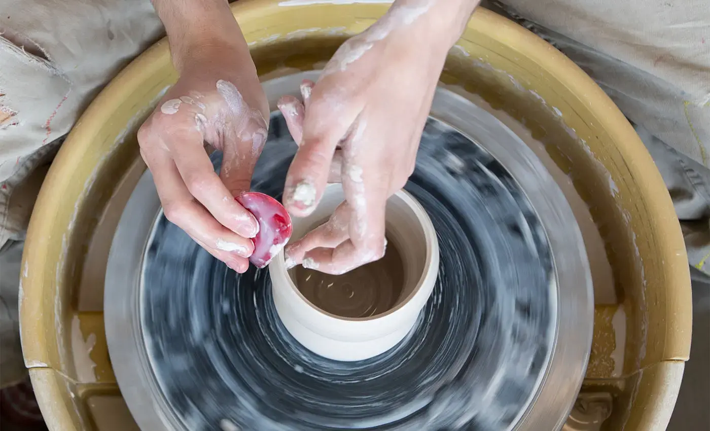 student's hands working with clay on a spinning potter's wheel