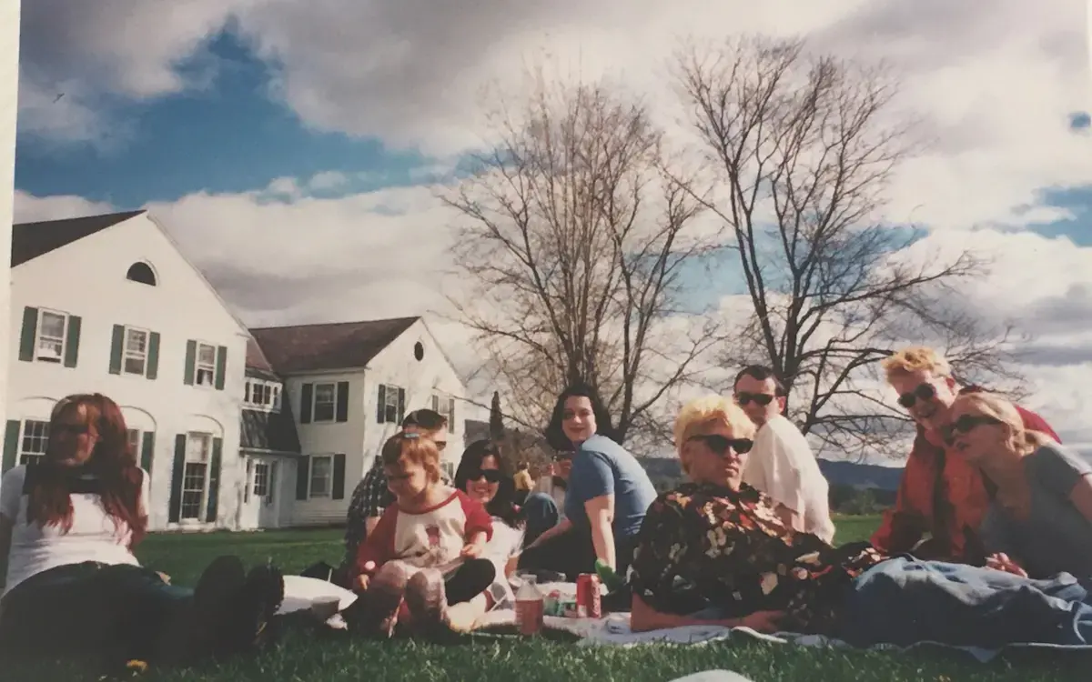 a group of students sit on a lawn with white houses in the background